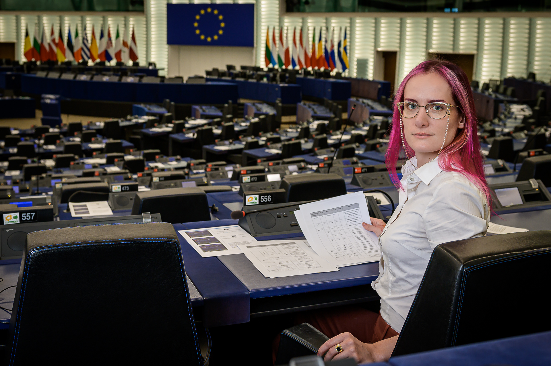 Markéta seating at the back of the empty plenary room, looking back at the camera